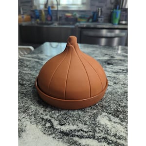 Pode incluir: Um recipiente de cerâmica marrom em forma de cebola com tampa. O recipiente tem uma base redonda e um topo bulboso com cristas verticais. A tampa tem um pequeno botão pontiagudo na parte superior. O recipiente está em uma bancada salpicada.