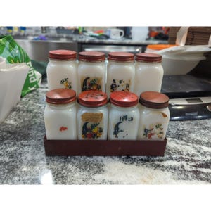 Puede incluir: Un juego de especiero vintage con ocho tarros de vidrio blanco. Cada tarro tiene una tapa de metal roja y una etiqueta decorativa. Los tarros están dispuestos en una bandeja de metal rojo oscuro, ideal para la cocina.
