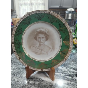 Op de afbeelding: Een ronde decoratieve keramische bord met een sepia-kleurig portret van koningin Elizabeth II met een kroon en ketting in het midden. De brede rand is smaragdgroen met ingewikkelde gouden filigraan bloemmotieven en een gouden rand.