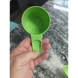 Puede incluir: Una taza medidora verde brillante de 3/4 de taza. La taza de plástico tiene un asa con el texto "3/4 CUP" impreso. La taza se sostiene en una mano. Perfecta para medir ingredientes.