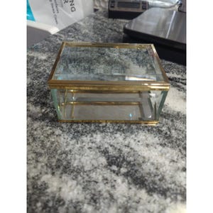 Può includere: Una scatola rettangolare di vetro con una cornice in metallo color oro. La scatola ha un design trasparente e trasparente, e anche il coperchio è in vetro. La scatola è su una superficie grigia maculata.