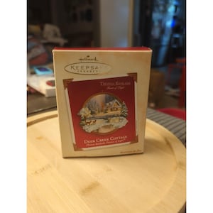 May include: A rectangular Hallmark Keepsake Ornament box featuring a red and cream design. The front displays a Thomas Kinkade painting titled Deer Creek Cottage, showing a snow-covered cabin, trees, and deer. The text reads Hallmark Keepsake Ornament.