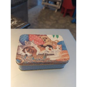 May include: A rectangular metal storage tin featuring a colorful illustration on the lid of a beagle puppy, a ginger tabby cat, and a small golden-colored puppy resting together on a woven rug against a background of blue and red patterned pillows.