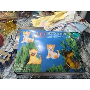 May include: A box of "Kitty Kats" patio-party lights. The box is blue with illustrations of orange and white cat figurines. The text on the box reads "Colorful Patio Lights for Festive Outdoor Lighting."