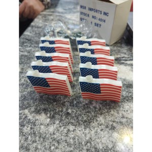 Puede incluir: Un conjunto de pequeñas banderas americanas, con rayas rojas y blancas y un rincón azul con estrellas. Las banderas están dispuestas en filas, con una base blanca y un pequeño cilindro blanco. La caja en el fondo dice "MSR Imports Inc".