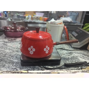 May include: A red enamel saucepan with a wooden handle and lid. White daisy patterns decorate the sides of the pot. The saucepan is sitting on a black scale.