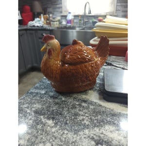 May include: A ceramic chicken-shaped container with a lid. The container is primarily brown with a red comb and yellow beak. The container is sitting on a gray countertop.
