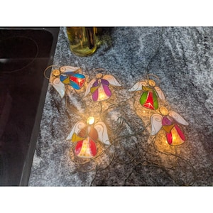 May include: A string of decorative lights featuring four angel-shaped ornaments with stained glass-style panels in red, blue, purple, green, and yellow. Each angel figure has white wings and a small trumpet, illuminated by internal warm white bulbs.