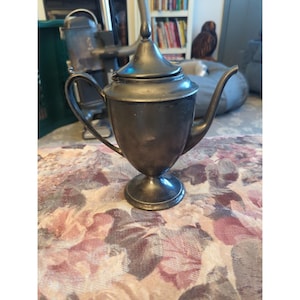 Op de afbeelding: Een kleine, antieke zilverkleurige theepot met een deksel en een gebogen tuit. De theepot heeft een handvat en staat op een voetstuk. De achtergrond omvat een tafelkleed met bloemenpatroon en een vage binnenkant.