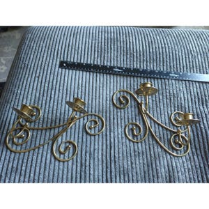 May include: Two gold-toned metal wall sconces featuring curved, scroll-like wire frames and two candle cups each. A black ruler is positioned above the sconces, showing a length of approximately 12 inches for the metal candle holder structures.