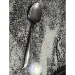 May include: A stainless steel spoon with a rounded bowl and a long, slender handle. The handle features a starburst design near the base. The spoon is set against a speckled gray and white surface.