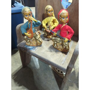 May include: Three vintage figurines of musicians in colorful outfits. Each doll is blonde with blue eyes and holds a musical instrument. The figurines are on a wooden chair with a distressed finish. The dolls are dressed in blue, yellow, and red.