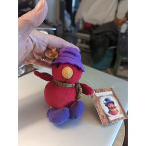 May include: A plush red bird toy wearing a purple hat with a small orange flower, a patterned scarf, and purple feet. The bird has a tan beak and black eyes. A rectangular cardboard tag is attached, featuring an illustration of the bird.