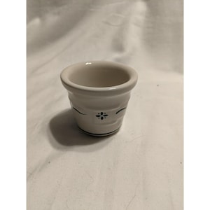 Peut inclure: Une petite tasse en céramique blanc cassé avec un bord évasé et un corps arrondi. L'extérieur présente un motif décoratif bleu foncé composé d'une fleur centrale à quatre pétales flanquée de lignes courbes et d'une fine bande horizontale.