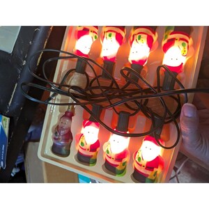 May include: A string of eight illuminated Santa Claus-shaped light bulbs connected by a black electrical cord, resting in a molded plastic tray. Each bulb features a red and white Santa figure with a glowing face, arranged in two rows of four.