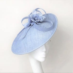 May include: A light blue, wide-brimmed hat with a floral accent. The hat features a large, circular brim and is adorned with a cluster of fabric flowers and decorative ribbons. The hat is displayed on a white mannequin head.