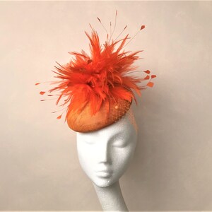 Könnte beinhalten: Ein leuchtend orangefarbener Fascinator mit einer großen Federboa und einer kleinen, runden Basis. Der Fascinator sitzt auf einem weißen Mannequin-Kopf.