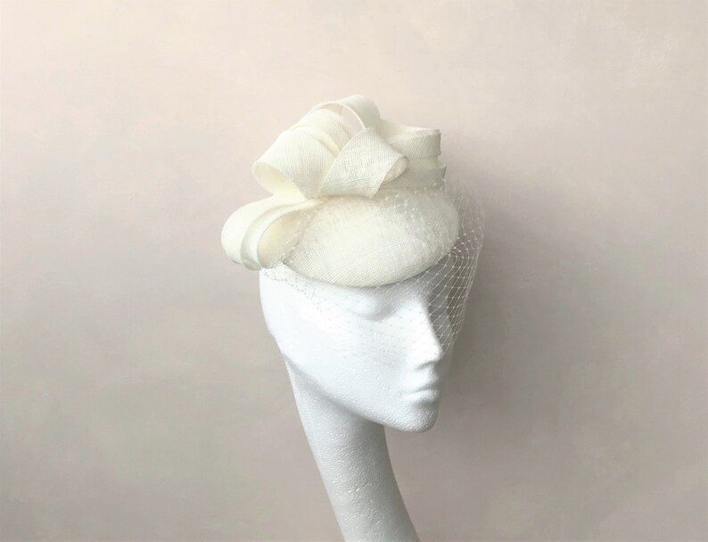 Puede incluir: Un sombrero de pastilla blanco con un gran lazo de tela blanco en la parte superior. El sombrero est&aacute; adornado con un velo blanco.
