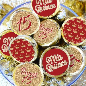 May include: A clear glass bowl filled with chocolate candies wrapped in gold foil and topped with red and gold round stickers. The stickers have various designs including the number 15, the words 'Sweet 15', 'Mis Quince', and a pattern of gold crowns on a red background.