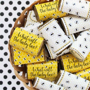 May include: A basket filled with small chocolate bars wrapped in yellow and white paper with a bee pattern. The wrappers have the text "What will the baby bee?" printed on them.