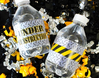 Puede incluir: Dos botellas de agua de plástico transparente con tapas blancas y etiquetas que dicen "UNDER CONSTRUCTION" en amarillo y negro. Las etiquetas tienen un diseño de placa de diamante. Las botellas están rodeadas de serpentinas metálicas amarillas, negras y plateadas.