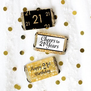 May include: Three gold and black chocolate bar wrappers with text. The first wrapper has the number "21" in gold on a black background. The second wrapper has the text "Cheers to 21 Years" in black on a gold background. The third wrapper has the text "Happy 21st Birthday" in black on a gold background.