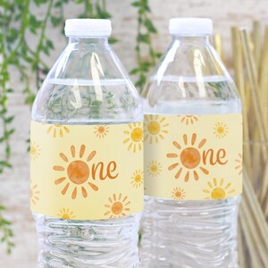 May include: Two clear plastic water bottles with yellow and orange labels. The labels have a watercolor sun design and the text "one".