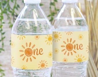 Puede incluir: Dos botellas de agua de plástico transparente con etiquetas amarillas y naranjas. Las etiquetas tienen un diseño de sol de acuarela y el texto "one".