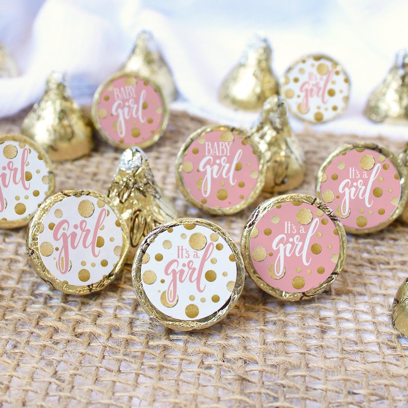 May include: Gold foil-wrapped chocolate candies with pink and gold "It's a girl" and "Baby Girl" stickers. The stickers have a pink background with gold polka dots and white text. The candies are arranged on a burlap surface.