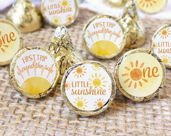 May include: Close-up of gold foil-wrapped chocolate candies with circular labels. The labels feature sun illustrations and phrases like "Little Sunshine" and "First Trip around the sun." The candies are arranged on a textured, tan surface.