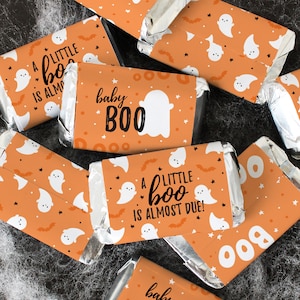 May include: Orange and white Halloween-themed chocolate bar wrappers with ghost and bat designs. The wrappers have text that reads "A little boo is almost due!" and "baby boo".