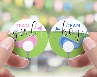 May include: Two circular gender reveal party favors. One has a pink golf club and the text "Team Girl". The other has a blue golf club and the text "Team Boy". Each favor features a golf ball on a tee and a small flag.