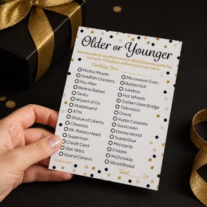 May include: A party game card with the text "Older or Younger" and a list of items to guess the age of. The card is held by a hand and is decorated with black and gold polka dots. A black gift box with a gold ribbon is in the background.