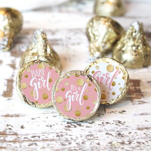 May include: Close-up of pink and gold baby shower candy. The candy wrappers say "Baby Girl" and "It's a girl" in white script. Gold polka dots are scattered across the pink wrappers. Several gold foil wrapped candies are in the background.