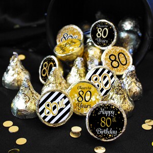 Puede incluir: Pegatinas redondas doradas y negras con el texto "Cheers to 80 Years", "Happy 80th Birthday" y "80th" sobre un fondo a rayas blanco y negro. Las pegatinas est&aacute;n colocadas en besos de chocolate.