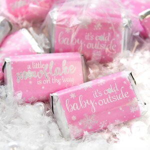 May include: Pink candy bars wrapped in silver foil, with white text and snowflake designs. The text on the wrappers reads "a little snowflake is on the way" and "baby it's cold outside". The candy is surrounded by white decorative fluff.