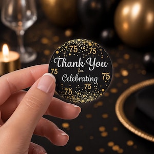 May include: A black and gold round sticker with the text "Thank You for Celebrating 75". The sticker has gold confetti and the number 75 in gold. The sticker is held by a person's hand.