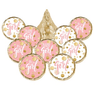 May include: A collection of chocolate candies with pink and white wrappers. The wrappers are decorated with gold polka dots and the phrases "It's a girl" and "Baby Girl" in pink script. A gold foil-wrapped chocolate kiss is in the center.