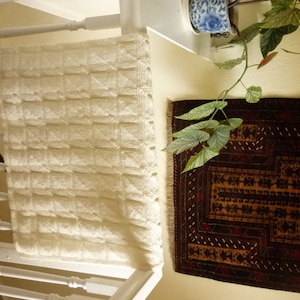 May include: A white crocheted blanket with a square pattern hangs over a white railing. The blanket is made of soft yarn and has a delicate texture.