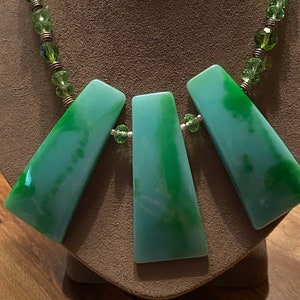 Könnte beinhalten: Eine grüne und blaue Halskette mit drei großen, dreieckigen Anhängern. Die Halskette besteht aus grünen Glasperlen und einer silbernen Kette.