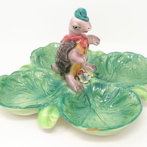 May include: A ceramic dish shaped like a four-leaf clover with a small turtle figurine on top. The dish is green and has a glazed finish. The turtle is wearing a blue hat and has a pink body, brown shell, and yellow accents.