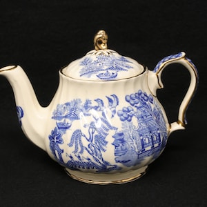 May include: Cream-colored teapot with a blue and white Willow pattern design. The teapot has a curved spout, a rounded body, and a lid with a gold finial. The handle and rim are also trimmed with gold.