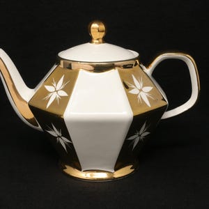 May include: A white and gold teapot with a geometric design. The teapot features a white body with gold accents and a gold lid. White floral designs are on the gold sections. The handle and spout are also gold.