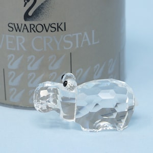 May include: A clear crystal hippopotamus figurine with black eyes. The figurine is sitting on a light blue surface. The box it came in is visible in the background. The box is silver and has the words "Swarovski Silver Crystal" printed on it.