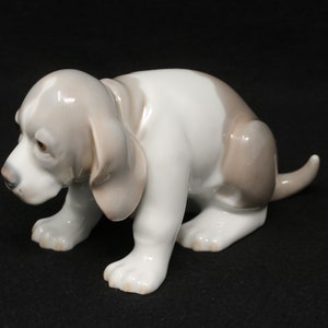 May include: A porcelain figurine of a white and grey basset hound puppy sitting with its head down. The puppy has brown eyes and a long, floppy tail.