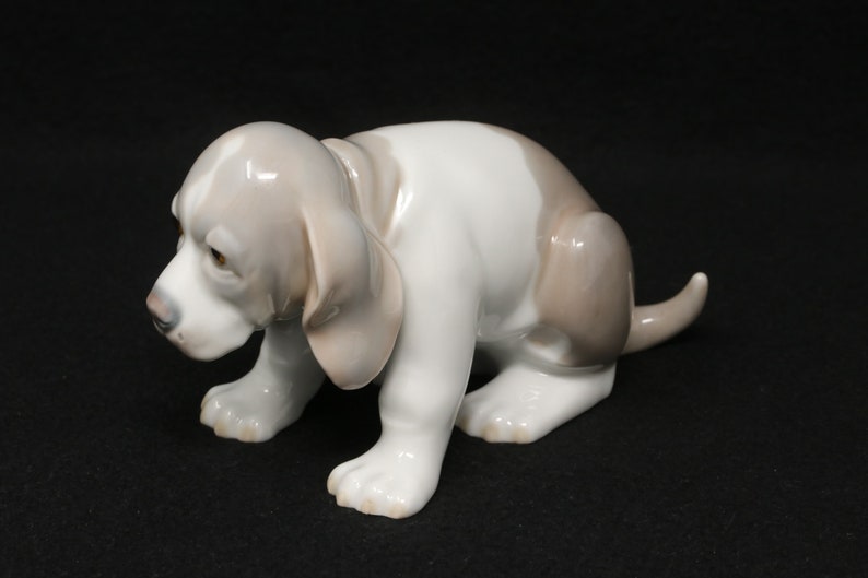 May include: A porcelain figurine of a white and grey basset hound puppy sitting with its head down. The puppy has brown eyes and a long, floppy tail.