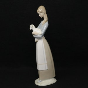 May include: A porcelain figurine of a woman holding a lamb. The woman is wearing a long, light blue sleeved top, a white apron, and a long, tan skirt. The lamb is white. The figurine is on a white base and set against a black background.