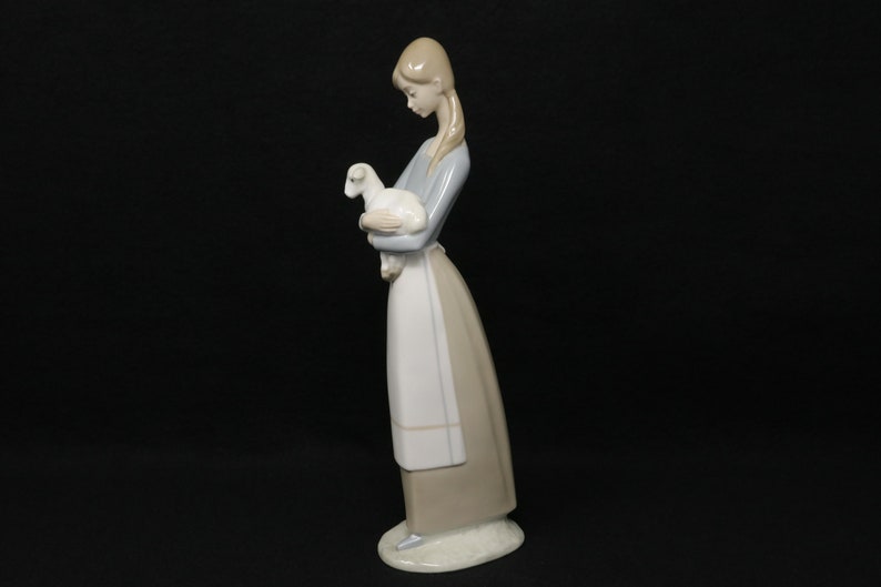 May include: A porcelain figurine of a woman holding a lamb. The woman is wearing a long, light blue sleeved top, a white apron, and a long, tan skirt. The lamb is white. The figurine is on a white base and set against a black background.
