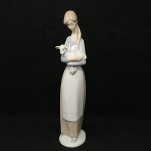 May include: A porcelain figurine of a woman holding a lamb. The woman is wearing a long, light-colored dress with a matching apron. The lamb is white with a light blue hue. The figurine is on a white circular base.
