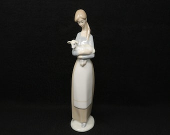 Könnte beinhalten: Porzellanfigur einer Frau, die ein Lamm hält. Die Frau trägt ein langes, helles Kleid mit passender Schürze. Das Lamm ist weiß mit einem hellblauen Farbton. Die Figur steht auf einem weißen, kreisförmigen Sockel.